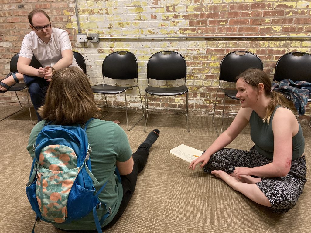 Two performers discussing on the ground with the director seated in a chair in the background.