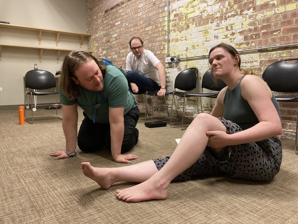 Two performers discussing on the ground with the director seated in a chair in the background.