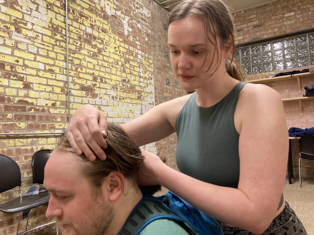 One performer is braiding the hair of another performer seated on the ground.