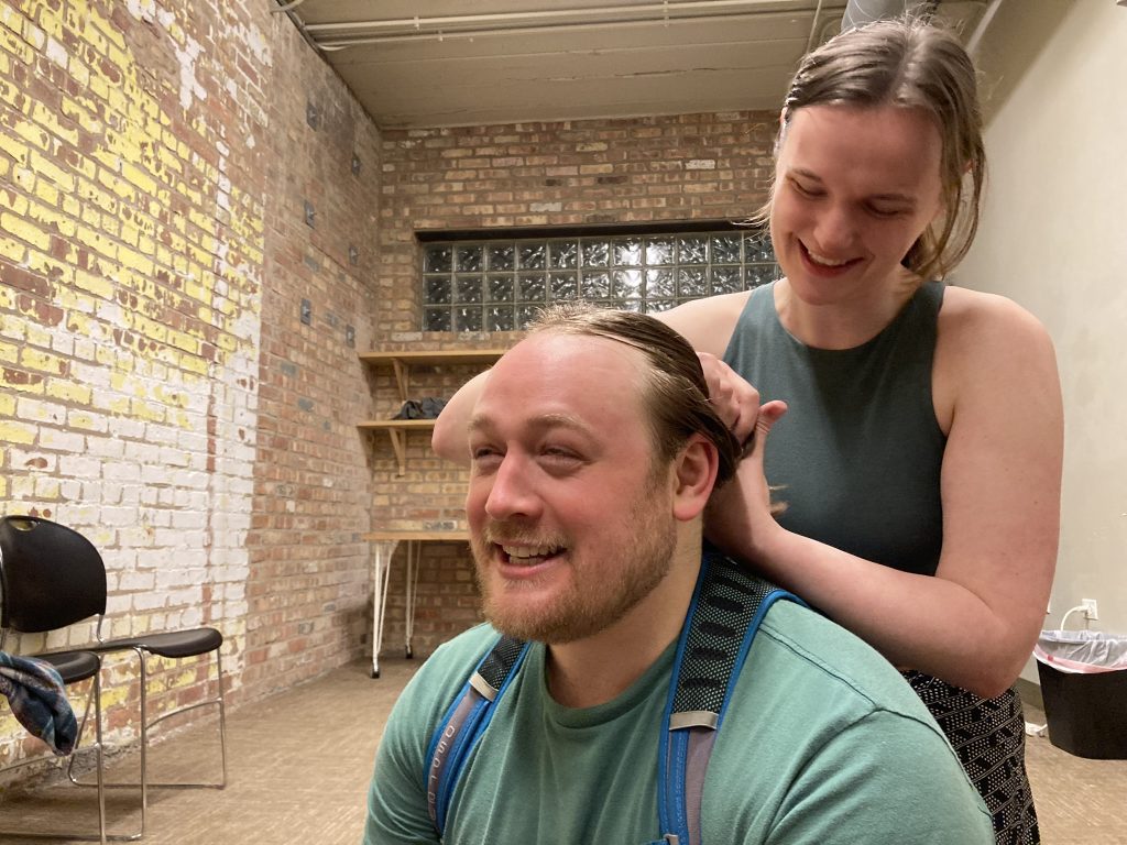 One performer is braiding the hair of another performer seated on the ground. They are laughing.