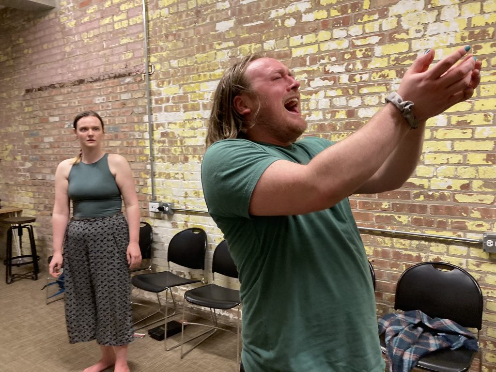 A performer is holding his hands together out in front of him and yelling while another is standing behind him in the background.