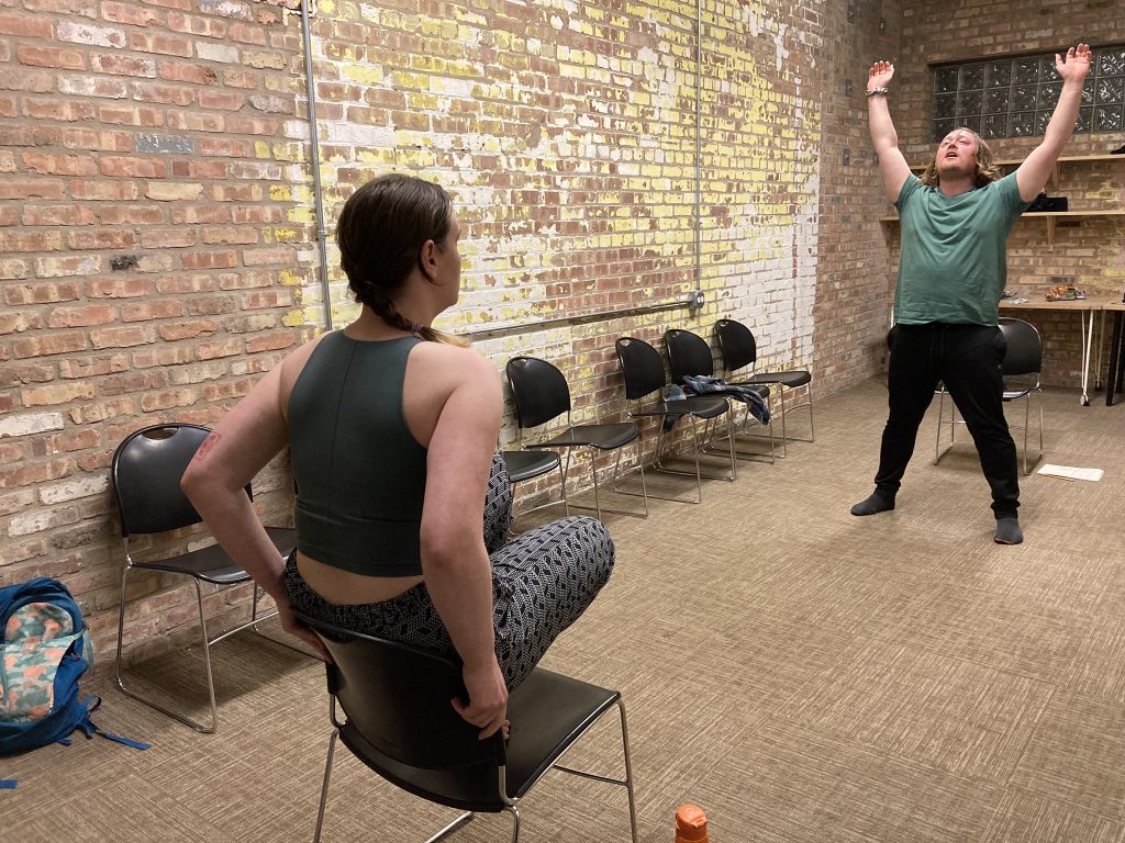 A performer is squatting on a chair while in front of them another performer is reaching their arms to the sky.