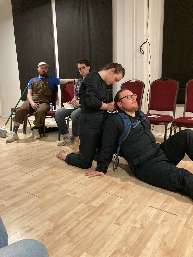 A performer is braiding another's hair on the ground while two audience members look on in the background.