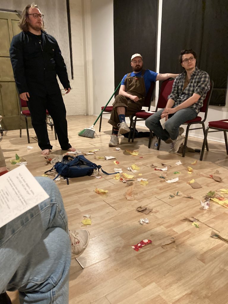 A performer stands above a backpack on the ground which is littered with pieces of trash. Two audience members look on in the background.