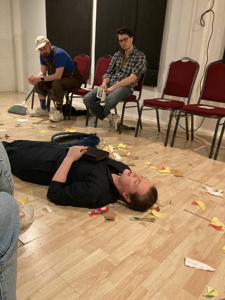 A performer lays on the ground holding a tape player while two audience members look on in the background.